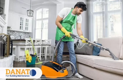 تنظيف السجاد في العين شخصًا يرتدي زي تنظيف وقفازات، وهو يستخدم جهاز تنظيف سجاد في غرفة معيشة حديثة. يجسد ذلك احترافية في تنظيف السجاد في العين باستخدام أدوات متقدمة لضمان نتائج ممتازة