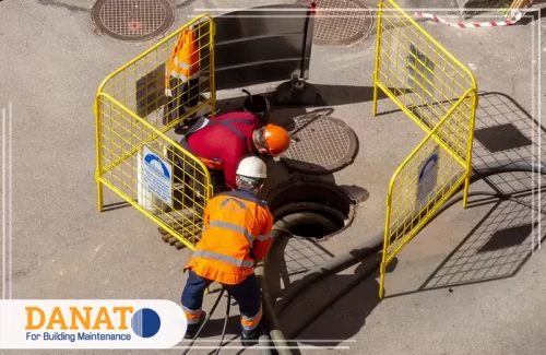 تسليك المجاري بالملح في دبي اثنين من الفنيين العاملين في مجال تسليك المجاري، يرتديان زي العمل مع خوذ، ويعملون على تسليك المجاري باستخدام أدوات متخصصة في مكان مغطى بالحواجز. تسليك المجاري بالملح في دبي يعد من الطرق الفعّالة