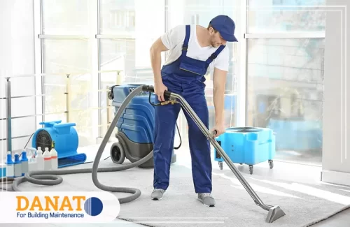 تنظيف السجاد على الناشف في دبي يظهر فني مختص من شركة دانات اللؤلؤ لصيانة المباني وهو يقوم بتنظيف سجاد باستخدام جهاز متطور للتنظيف الجاف داخل منزل حديث ومضاء طبيعياً. تُبرز الصورة خدمة تنظيف السجاد على الناشف في دبي