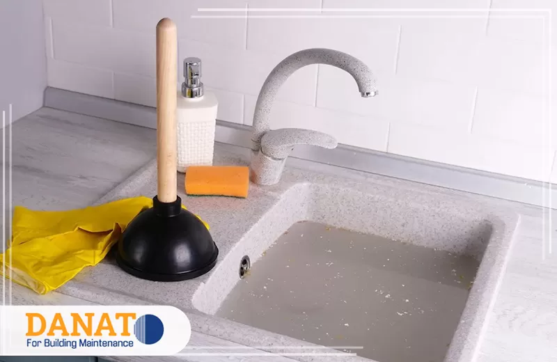 معالجة روائح الرطوبة والعفن في الشارقة 0545248958 دانات اللؤلؤ حوض مطبخ ممتلئ بمياه راكدة نتيجة انسداد الصرف، مع أدوات التنظيف المستخدمة لتسليك المواسير والحد من الروائح الكريهة.
